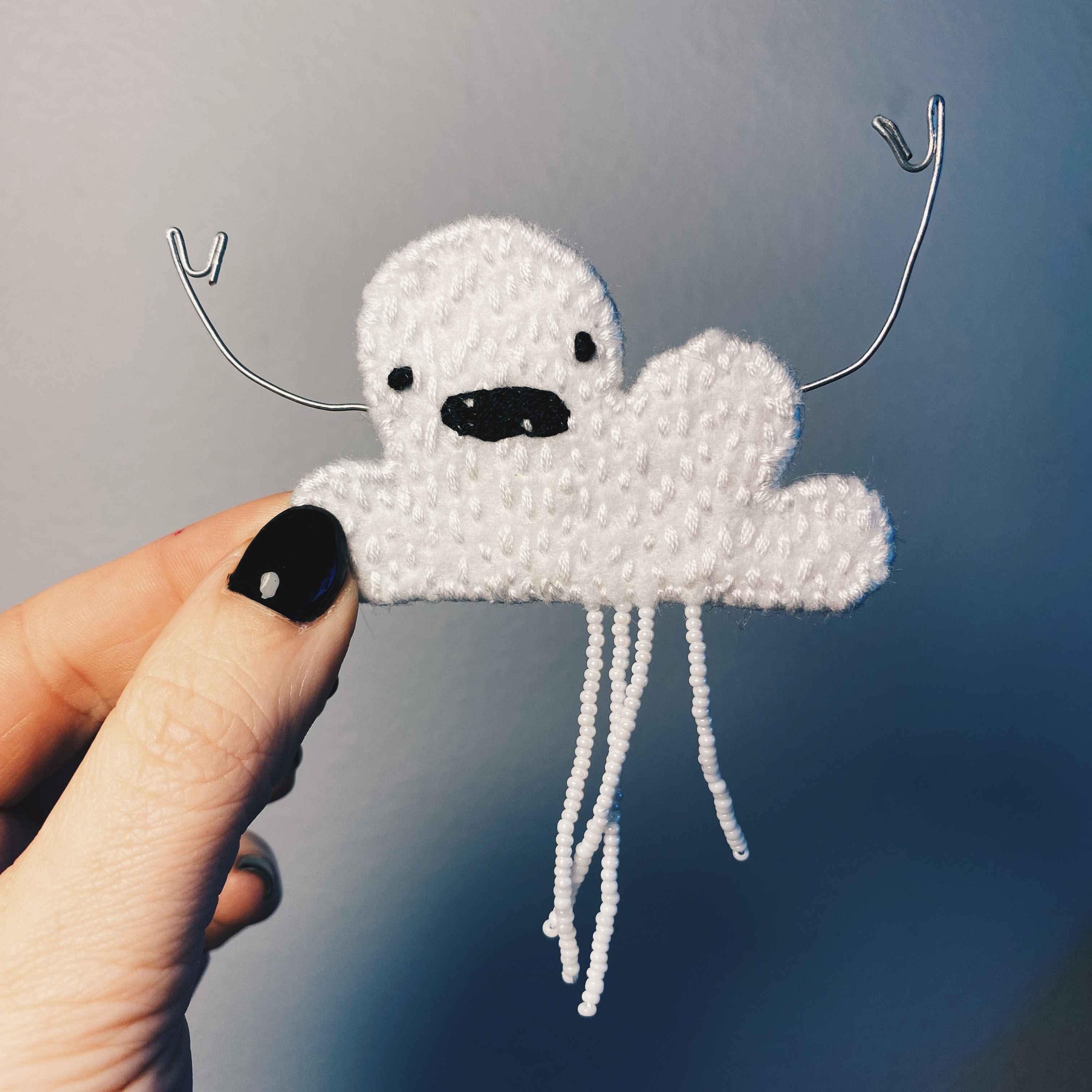 small embroidered cloud with face, held in hand with wire arms and beaded white rain fringe.