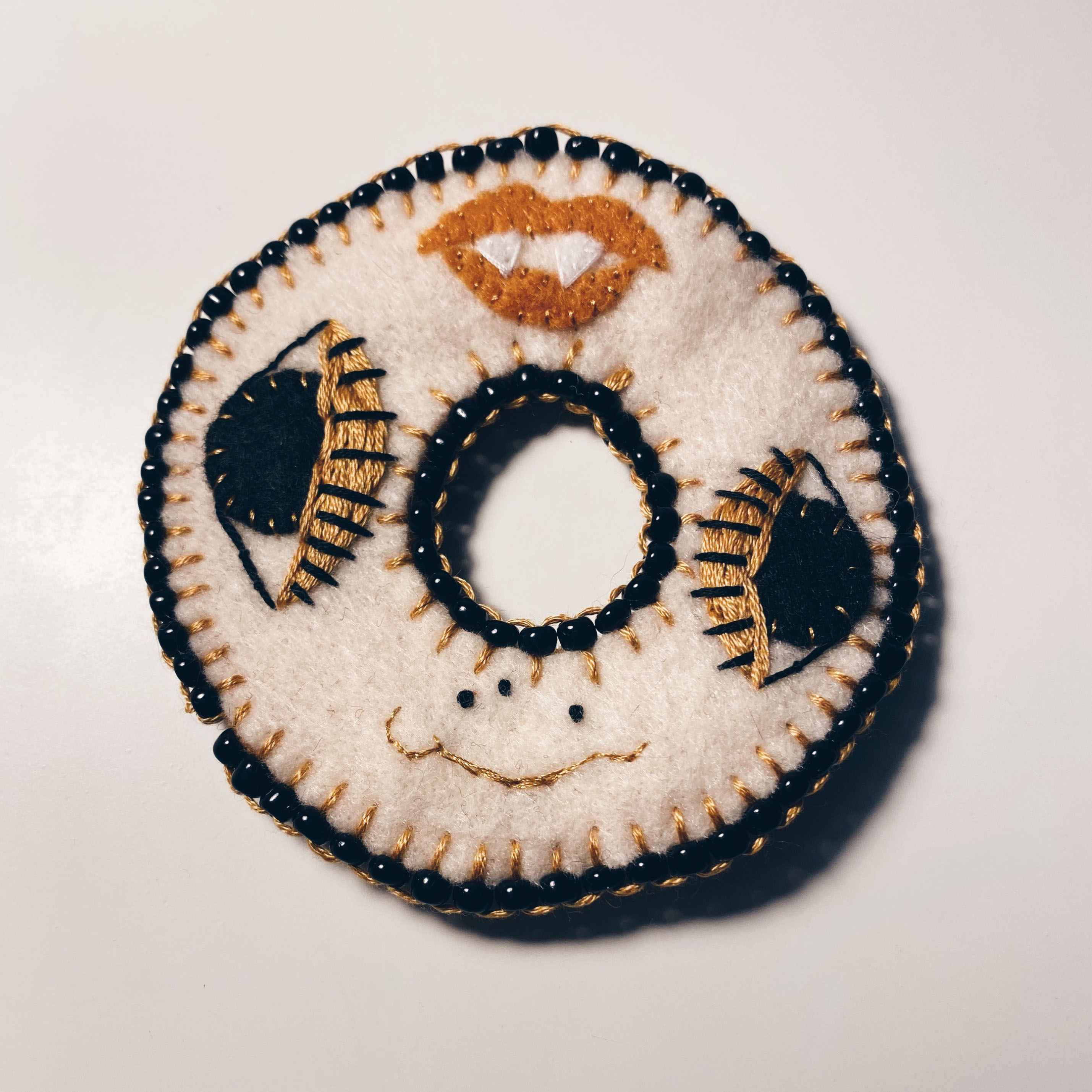 embroidered donut with black beads around the edge and tan face embroidered with vampire teeth lips, two eyes and nose going around the circle.