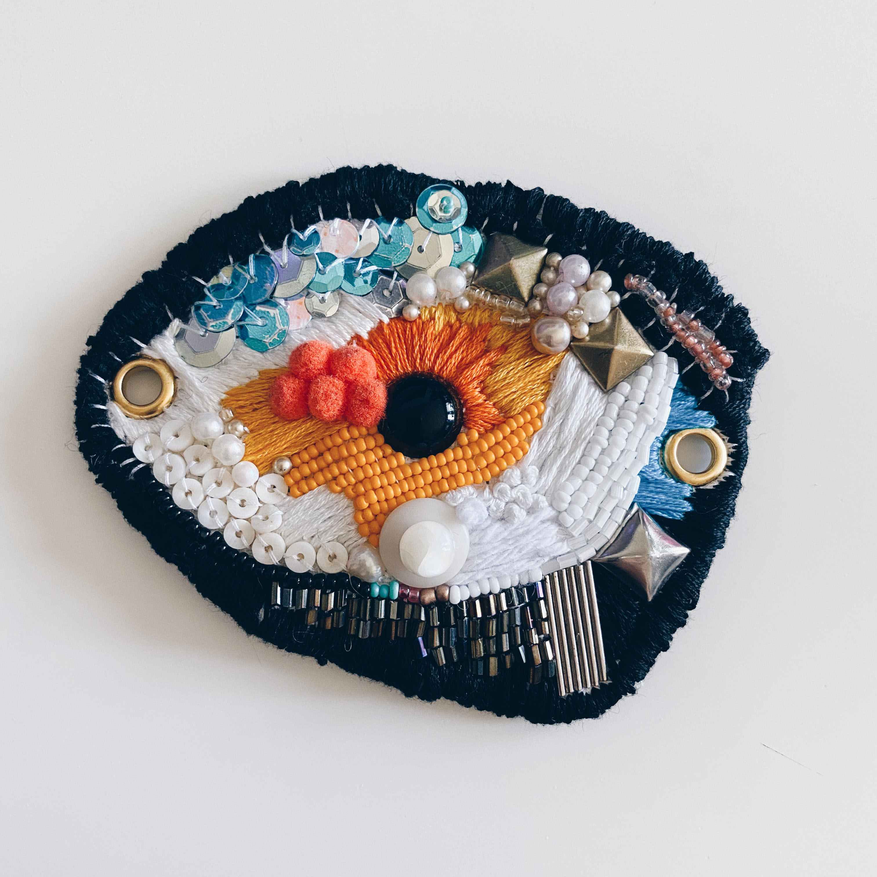 Large beaded embroidered eye with mixed materials in orange white and black.