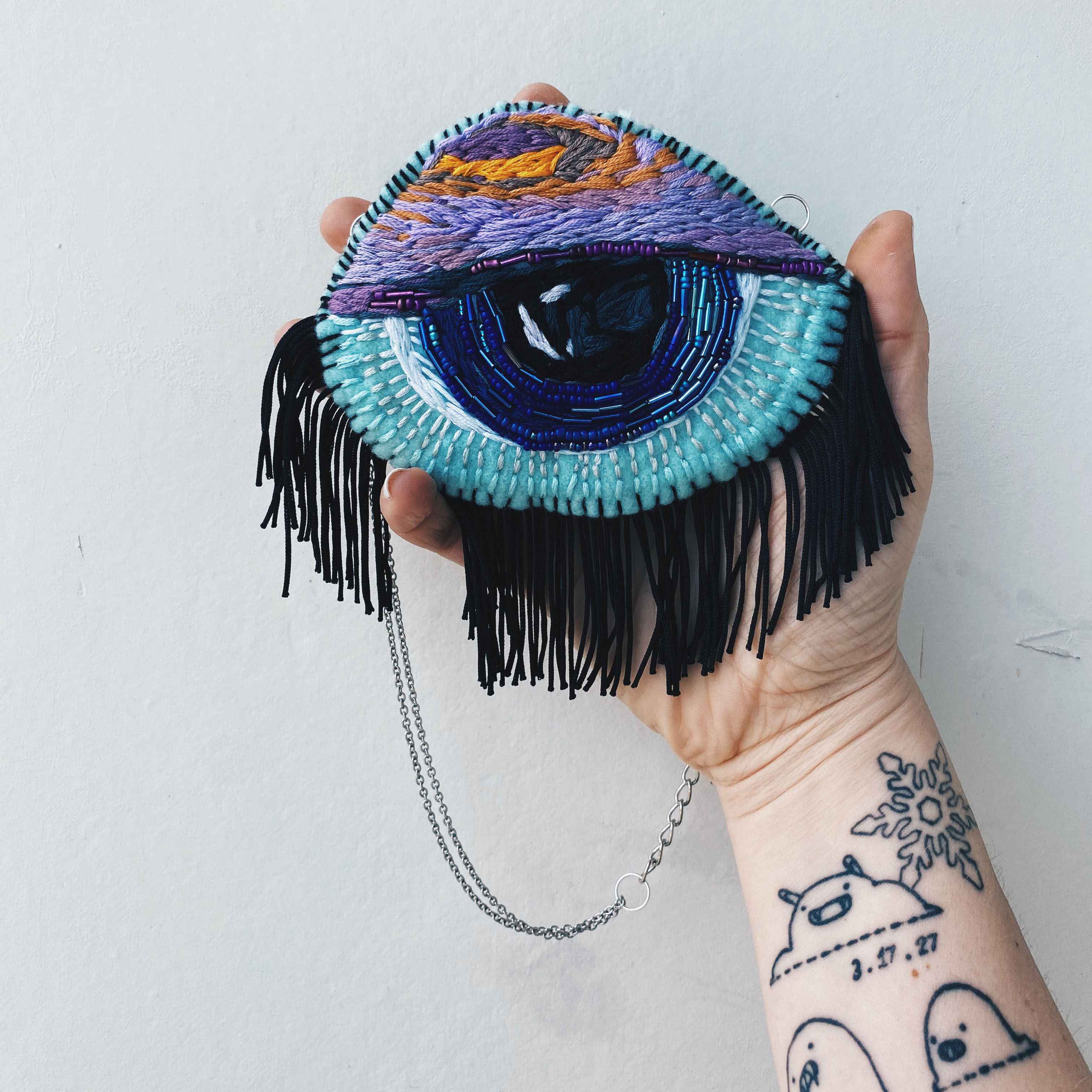handmade blue and purple beaded eye held in hand for scale. with fringe and chain hanging down.