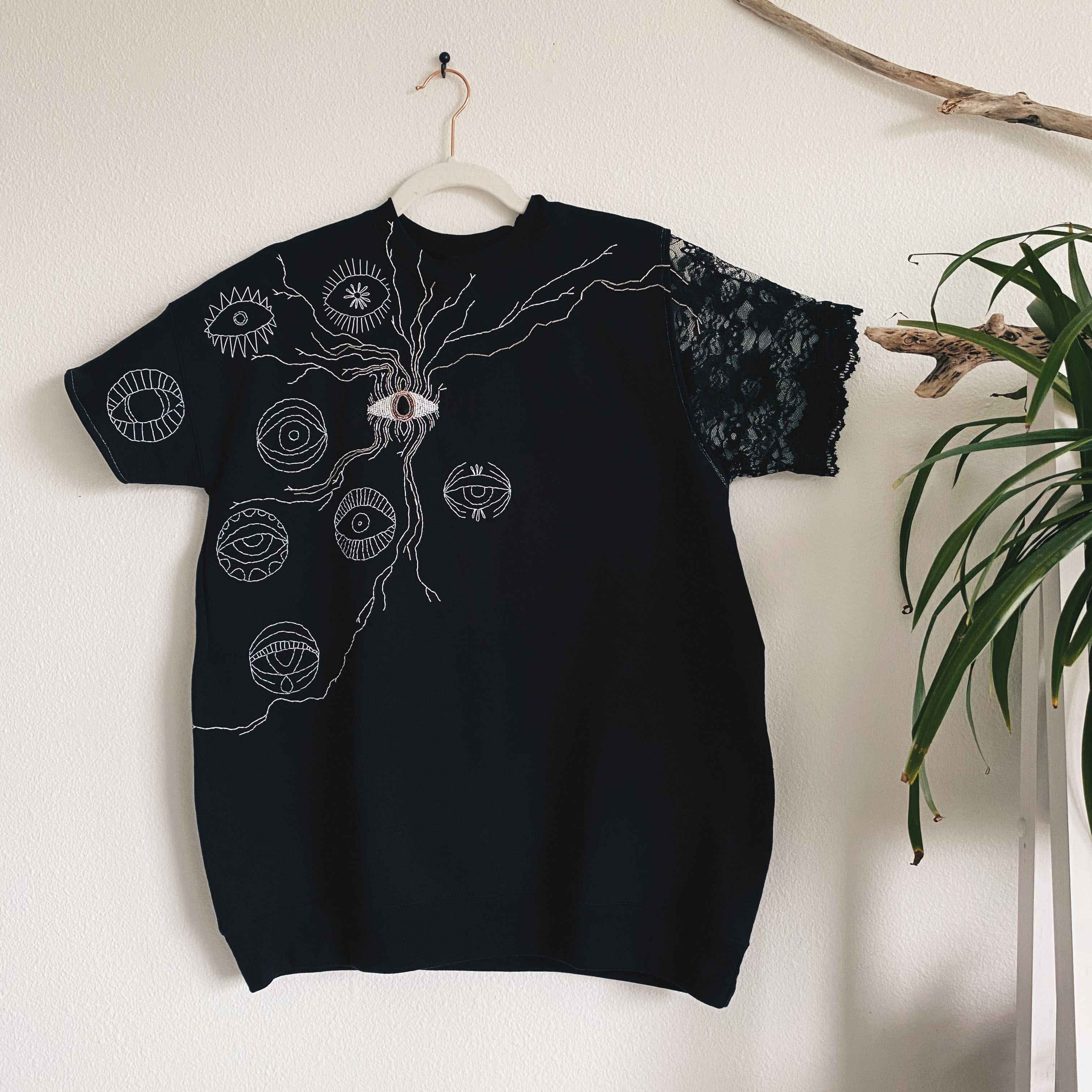 Black short sleeve sweater with hand embroidered and beaded eyes. shown on hanger on wall next to plants.